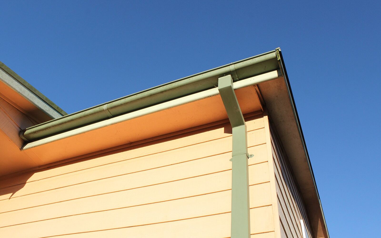 A metal rain gutter and downspout at the eave of a house against a blue sky